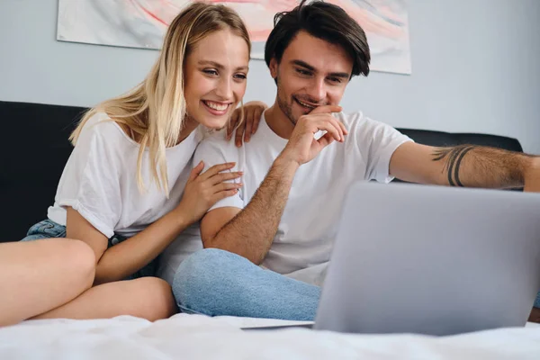 Schöne lächelnde blonde Frau und attraktive brünette Mann freudig Film auf Laptop zusammen. junges, fröhliches Paar in weißen T-Shirts sitzt zu Hause auf dem Bett — Stockfoto