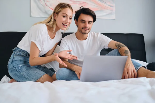 Fröhliche blonde Frau und lächelnder brünetter Mann, die fröhlich zusammen am Laptop arbeiten. junges schönes Paar in weißen T-Shirts sitzt zu Hause auf dem Bett — Stockfoto