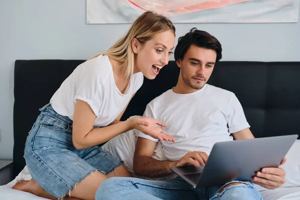 Szép érzelmi szőke nő vidáman látszó-ban laptop, míg vonzó barna ember dolgozik rajta. Fiatal szép pár fehér pólók ülnek együtt ágyban otthonos otthon — Stock Fotó