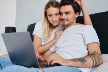 O dizüstü bilgisayar üzerinde çalışırken güzel sarışın kadın hayalle gülümseyen esmer adam sarılma. Evde birlikte yatakta oturan beyaz t-shirt genç çekici çift