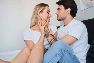 Yakışıklı esmer adam güzel gülümseyen sarışın kadının yüzüne yakın el tutarak ve hayali ona bakıyor. Modern rahat otelde birlikte yatakta oturan beyaz t-shirt genç güzel çift