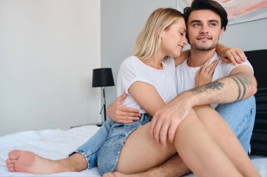 Güzel sarışın kadın mutlu ona sarılma ise Çekici gülümseyen esmer adam hayaldi kenara bakıyor. Modern otelde birlikte yatakta oturan beyaz t-shirt genç güzel çift