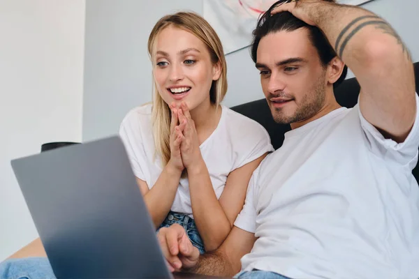 Junge brünette Mann und hübsche lächelnde blonde Frau in weißem T-Shirt fröhlich mit Laptop zusammen sitzen auf dem Bett in modernen gemütlichen Hotel — Stockfoto