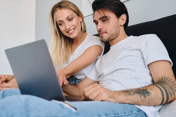 Young brunette man and beautiful smiling blond woman in white T-shirts happily using laptop together sitting on bed at cozy home — Stock Photo, Image