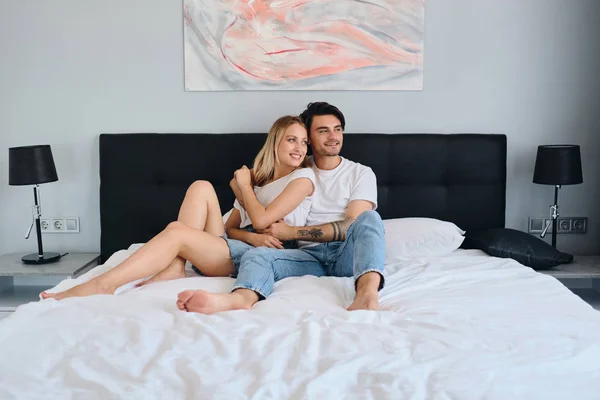 Beautiful blond woman and handsome brunette man hugging each other. Young attractive couple in white T-shirts lying on bed in modern cozy hotel happily looking aside together — Stock Photo, Image
