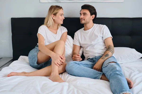 Jonge aantrekkelijke brunette man en mooie glimlachende blonde vrouw in witte T-shirts praten en dromerige naar elkaar kijken op bed in modern gezellig hotel — Stockfoto