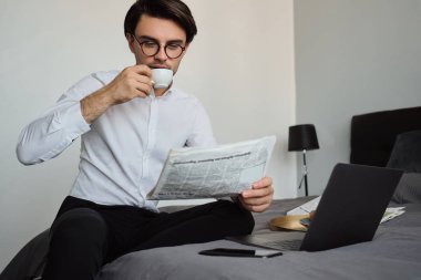 Beyaz gömlek ve gözlük genç yakışıklı esmer adam hayalkahve içme ve modern rahat otelde yakın dizüstü bilgisayar ve cep telefonu ile yatakta oturan gazete okuma