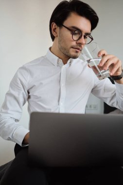 Beyaz gömlek ve gözlük genç güzel esmer adam rüya dizüstü bilgisayar üzerinde çalışan yatakta oturan ve modern otelde saf su içme