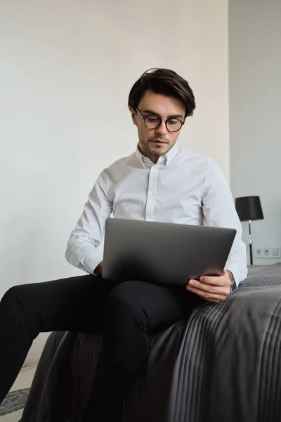 Joven hombre guapo morena en camisa blanca y anteojos sentados en la cama trabajando pensativamente en el ordenador portátil en el hotel moderno — Foto de Stock