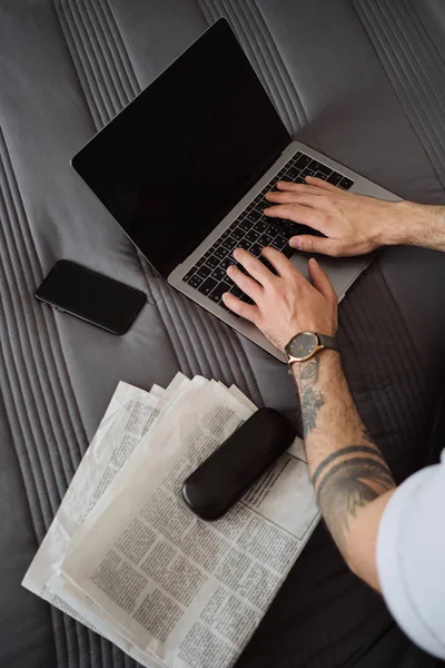Close up foto de mãos de homem com tatuagem trabalhando no laptop com celular, jornais e estojo para óculos perto na cama em casa — Fotografia de Stock