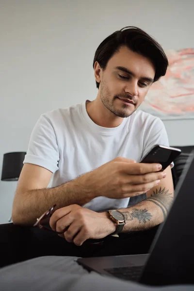 Joven atractivo morena sonriente hombre en camiseta blanca con gafas en la mano de ensueño utilizando el teléfono celular sentado en la cama con el ordenador portátil en el hogar moderno — Foto de Stock