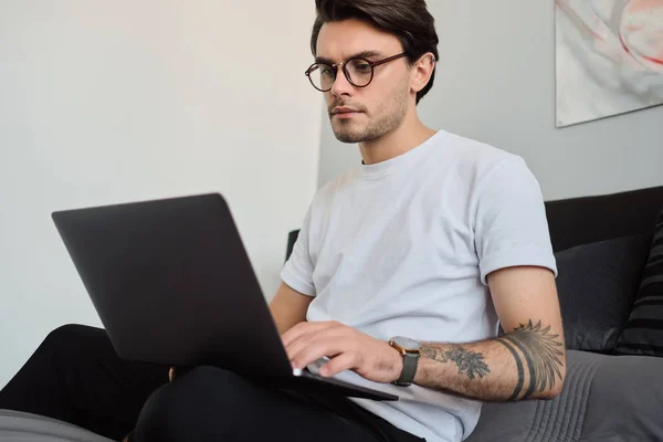 Joven morena atractiva hombre en camiseta y anteojos sentado en la cama en el hogar moderno cuidadosamente usando en el ordenador portátil — Foto de Stock
