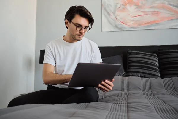 Joven hombre guapo morena en camiseta y anteojos sentado en la cama grande en el hogar moderno cuidadosamente trabajando solo en el ordenador portátil — Foto de Stock