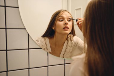 Beyaz elbiseli genç çekici kadın düşünceli banyoda maskara uygulayarak ayna yakınında duruyor