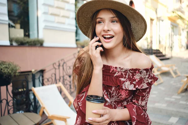 Young beautiful woman in dress and straw hat holding in hand cup of coffee to go and talking on celphone happily looking aside on cozy city street — Stock Photo, Image