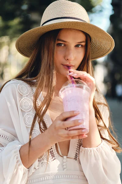 Junge schöne Frau in weißem Kleid und Hut, die Smoothie trinkt, schaut nachdenklich in die Kamera auf der gemütlichen Stadtstraße — Stockfoto