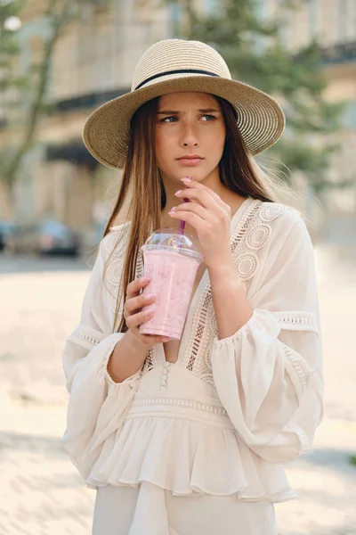 Jovem atraente chateado mulher em vestido branco e chapéu segurando smoothie para ir na mão, infelizmente, olhando de lado em pé na rua da cidade sozinho — Fotografia de Stock