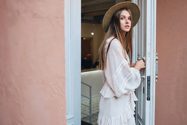 Fiatal gyönyörű nő, fehér ruhában és kalapban elgondolkodva keresi a kamera nyitó fehér ajtó a kávézó — Stock Fotó