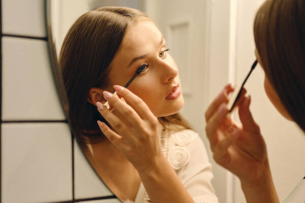 Close up young beautiful woman in white dress standing near mirror thoughtfully applying mascara in bathroom