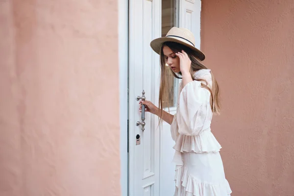 Young beautiful woman in white dress and straw hat thoughtfully looking down standing near white door on street