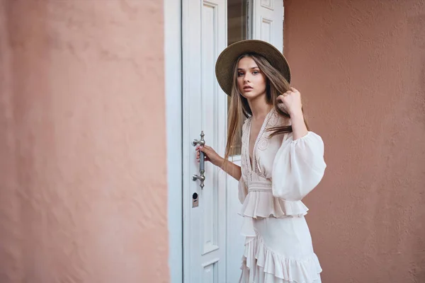 Jeune femme attrayante en robe blanche et chapeau de paille regardant de rêve dans la caméra debout près de la porte blanche sur la rue — Photo