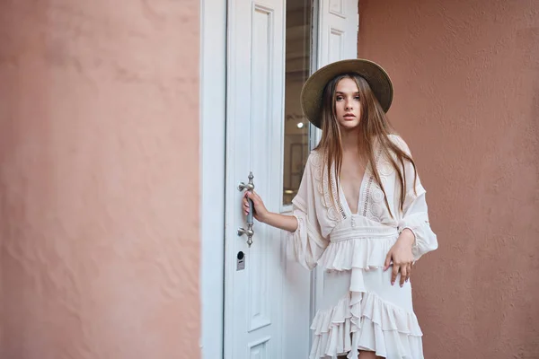 Jeune belle femme en robe blanche et chapeau de paille regardant attentivement dans la caméra debout près de la porte blanche sur la rue — Photo