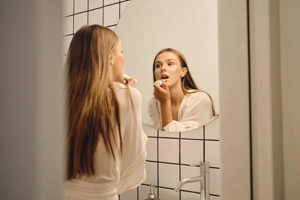 Молодая красивая женщина в белом платье мечтательно глядя в зеркало нанося помаду в ванной комнате — стоковое фото