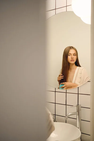 Jovem bela mulher séria em vestido branco pensativo olhando para o lado escovando o cabelo perto do espelho no banheiro — Fotografia de Stock