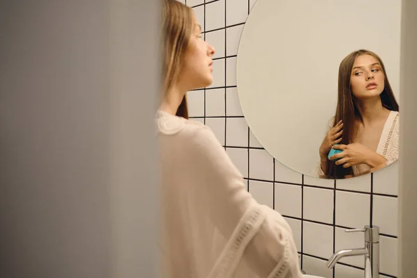 Nuori kaunis mietteliäs nainen valkoisessa mekossa huomaavaisesti sivuun harjaamalla hiuksiaan lähellä peiliä kylpyhuoneessa — kuvapankkivalokuva