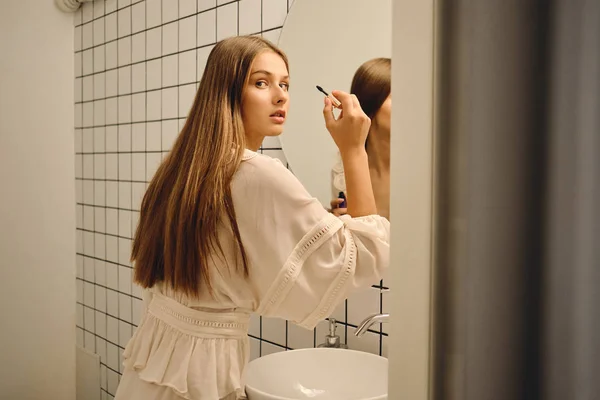 Mladá krásná žena v bílých šatech, stojící blízko zrcadla a zamyšleně se dívala do kamery v koupelně — Stock fotografie