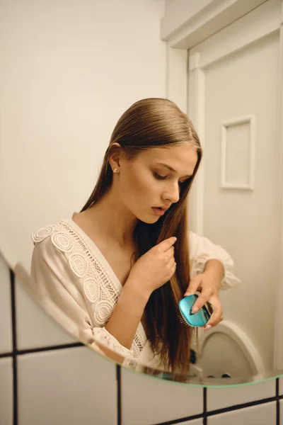 Jovem bela mulher pensativa em vestido branco de pé perto do espelho sonhadoramente escovando seu cabelo no banheiro — Fotografia de Stock