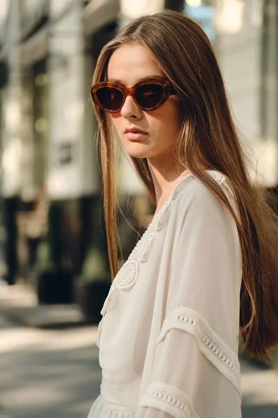 Fiatal gyönyörű nő napszemüveg és fehér ruhában elgondolkodva keresi a kamera állt a hangulatos városi utcában egyedül — Stock Fotó