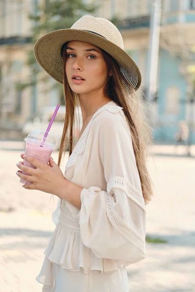 Young beautiful woman in white dress and hat holding smoothie to go in hand dreamily looking in camera standing on city street alone