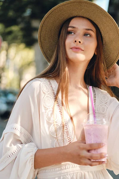 Jovem mulher bonita em vestido branco e chapéu segurando smoothie para ir na mão sonhadoramente olhando de lado em pé na acolhedora rua da cidade — Fotografia de Stock