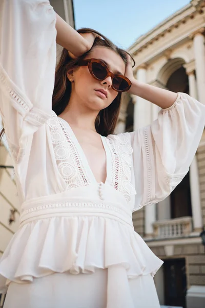 Fiatal gyönyörű nő napszemüveg és fehér ruhában elgondolkodva keresi a kamera kezében kezét fejjel állva a város utcában — Stock Fotó