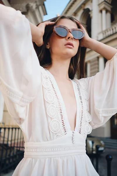 Молодая привлекательная женщина в солнечных очках и красивом белом платье вдумчиво глядя в сторону стоя на городской улице — стоковое фото