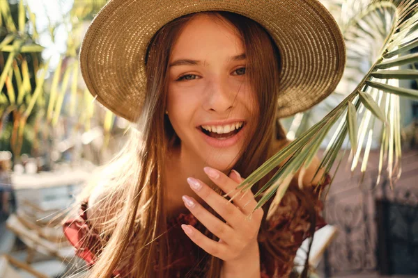 Gros plan jeune jolie femme souriante en robe et chapeau de paille regardant joyeusement dans la caméra avec de grandes feuilles vertes sur le fond dans un café de rue confortable — Photo