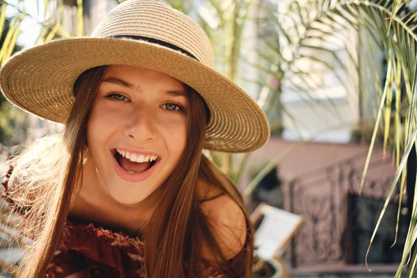 Close up young cheerful woman in dress and straw hat happily looking in camera with big green leaves on background in cozy street cafe — Stock Photo, Image