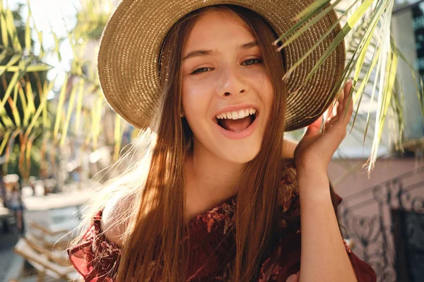 Close up young pretty cheerful woman in dress and straw hat happily looking in camera with big green leaves on background in cozy street cafe — Stock Photo, Image