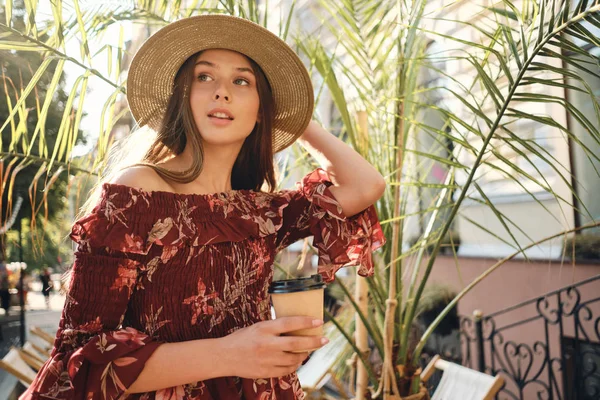 Young beautiful woman in dress and straw hat holding cup of coffee to go in hand dreamily looking aside with big green leaves on background in cozy street cafe — Stock Photo, Image