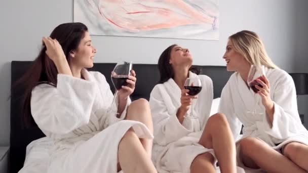 跟踪拍摄美丽的女孩穿着白色浴袍愉快地交谈和喝红酒在床上在酒店房间 — 图库视频影像
