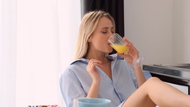 跟踪拍摄美丽的微笑的金发女郎在男性衬衫吃羊角面包享受味道和喝橙汁在现代厨房 — 图库视频影像