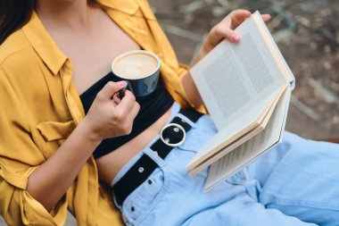Şehir parkında elinde kahve ve kitap bir fincan tutan sarı gömlek ve kot pantolon kadın kapatın