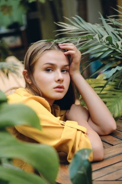 Genç çekici ciddi kahverengi saçlı kadın sarı gömlek li yandan düşünceli parkta yeşil yaprakları ile kamera bakarak baş eğilerek