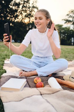Pretty gülümseyen genç kız mutlu şehir parkı piknik cep telefonu görüntülü sohbet konuşurken kulaklık ile ekose oturan