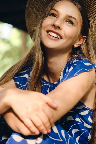 Portrait of beautiful cheerful girl in blue dress and hat happily sitting in city park — Stock Photo, Image