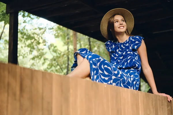 Jeune femme souriante attrayante en robe bleue et chapeau joyeusement regarder de côté dans le parc de la ville — Photo