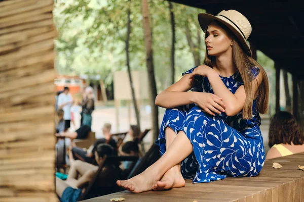 Jeune femme rêveuse attrayante en robe bleue et chapeau pieds nus regardant pensivement de côté dans le parc de la ville — Photo