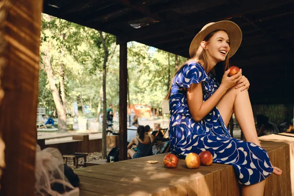 Jonge vrolijke vrouw in blauwe jurk en hoed Barefoot eten perzik gelukkig kijken opzij zittend op houten hek in stadspark — Stockfoto