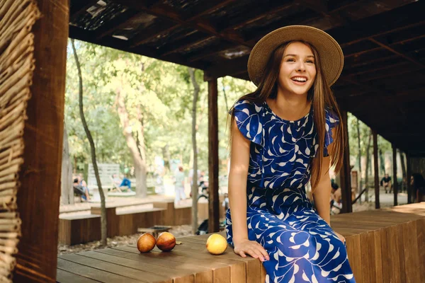 青いドレスと帽子をかぶった若いゴージャスな笑顔の女性は、市内の公園の木製のフェンスに美味しい桃と一緒に楽しく座っています — ストック写真